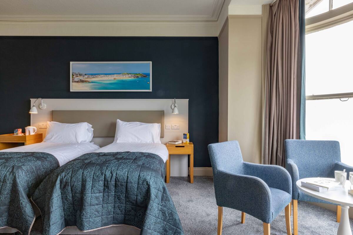 Habitación de hotel con dos camas, un cuadro de una playa, una ventana con cortinas y una pequeña mesa con sillas.