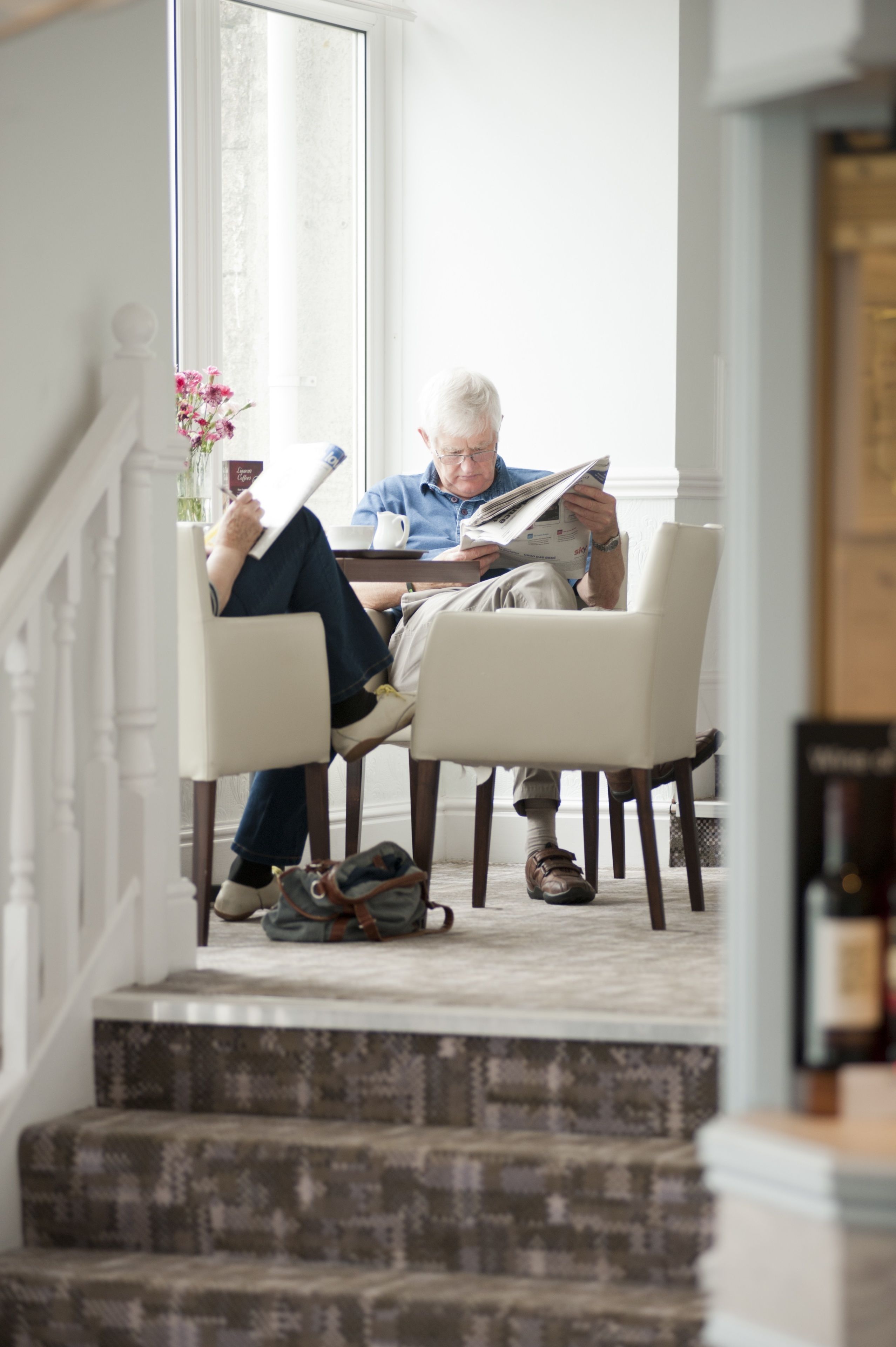 A person sitting at a table reading a newspaper in a well-lit room with a large window, a staircase, and a vase with flowers.