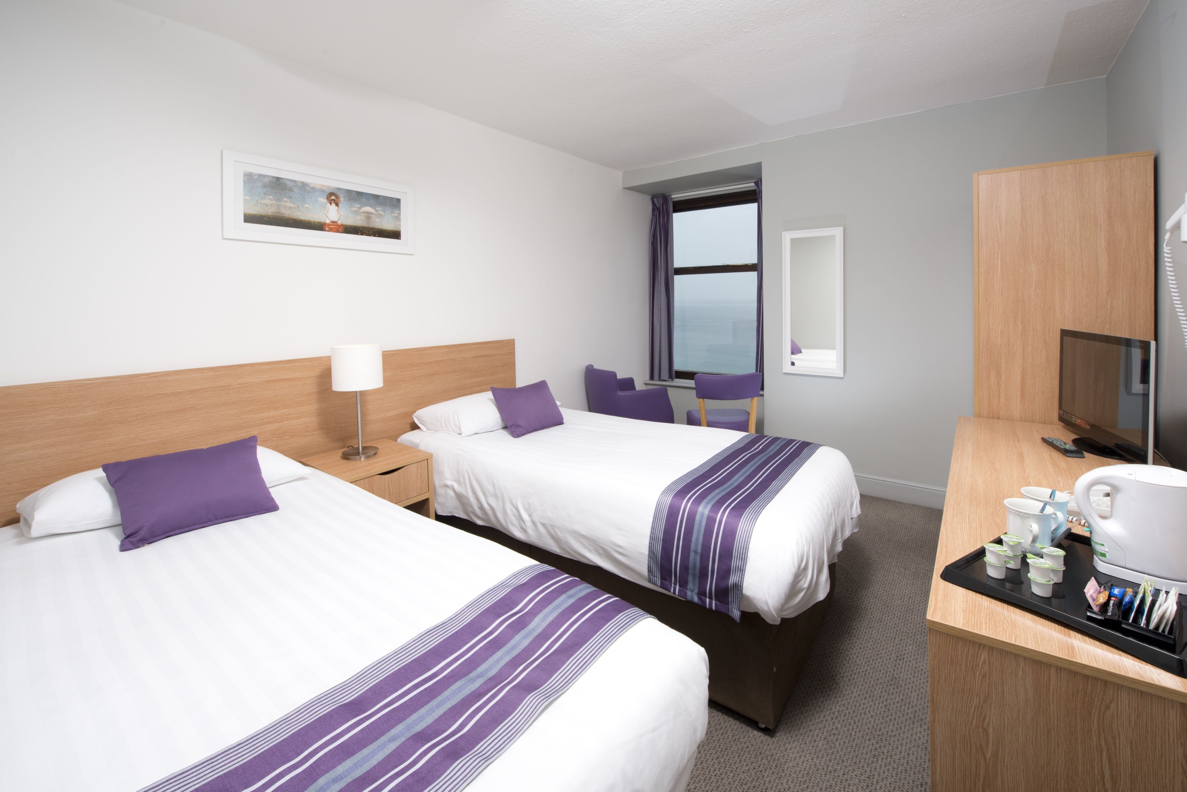 Een hotelkamer met twee bedden, een bureau met koffiezetapparaat en uitzicht op zee door een raam.