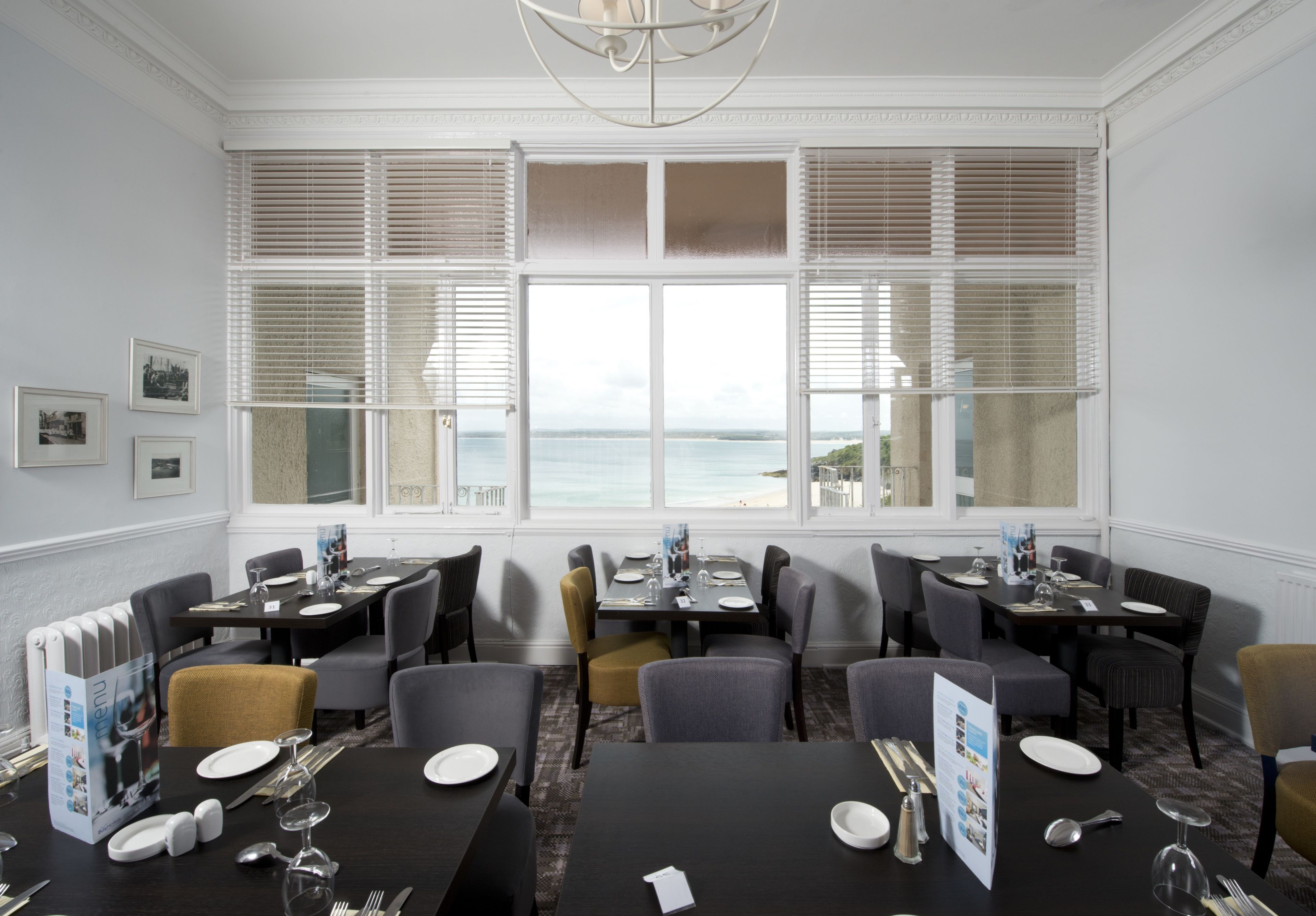 A dining area with tables set for four, large windows with blinds, and a view of the sea.