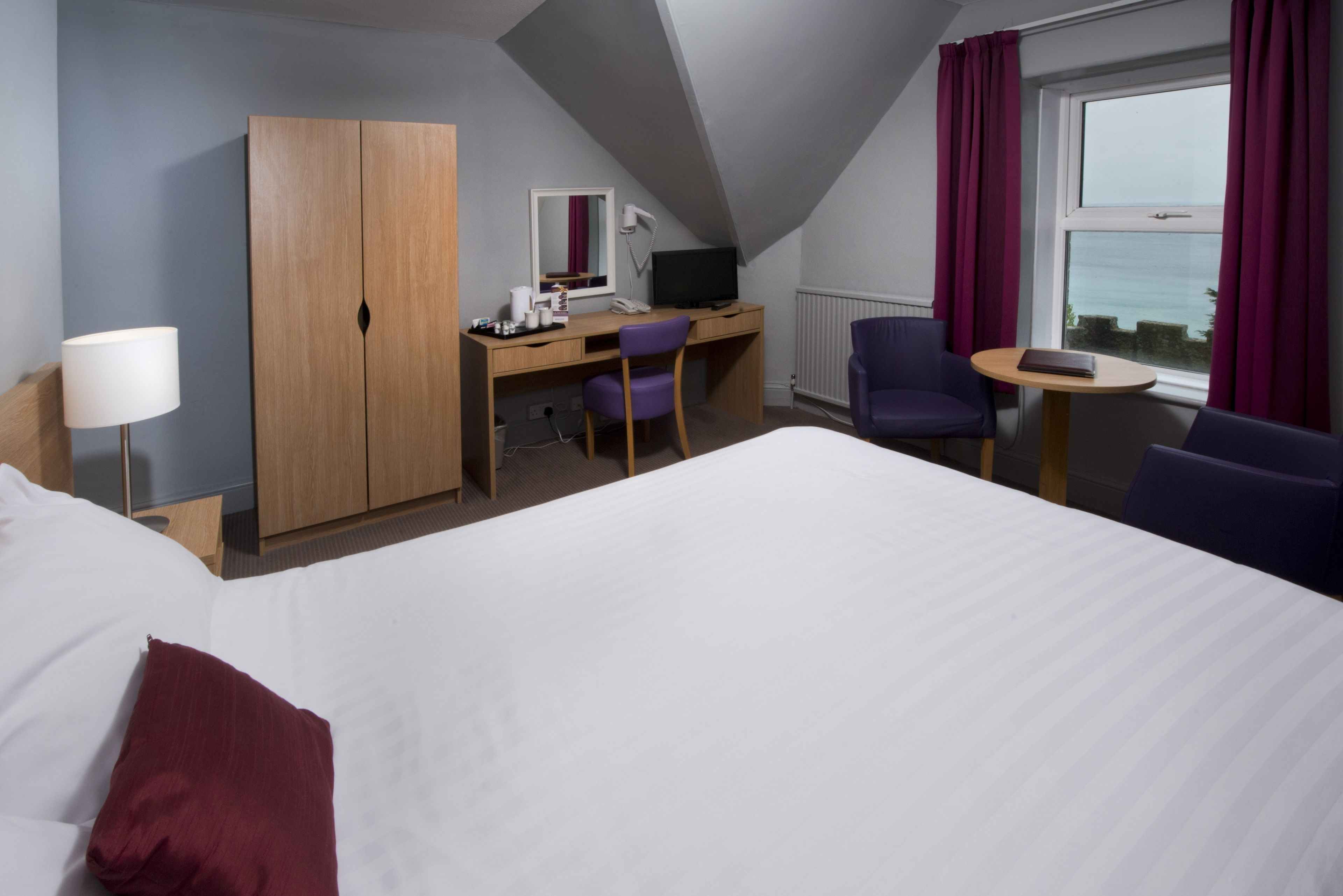 A hotel room with a large bed, a desk with a computer, two chairs, a small table, a lamp, and a view of the sea through a window.