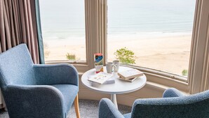 Una habitación con vista a la playa, que incluye dos sillas azules, una mesita con un libro y una taza, y una ventana con cortinas.