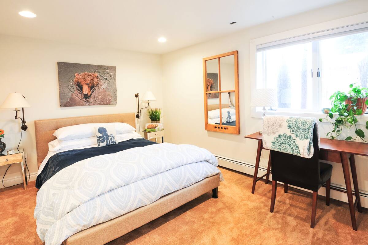 A neatly arranged bedroom with a bed, bedside tables, a mirror, a chair, and a window with a view.