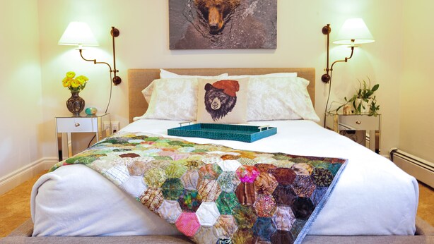 A neatly made bed with a quilt, two bedside lamps, a vase with flowers, and a bear-themed pillow. - Cranky Crow Bed and Breakfast (Kodiak)
