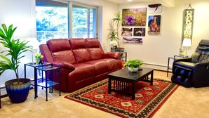 A living room with a red leather sofa, a small coffee table, a blue side table, a black massage chair, and a red and black patterned rug. - Cranky Crow Bed and Breakfast (Kodiak)