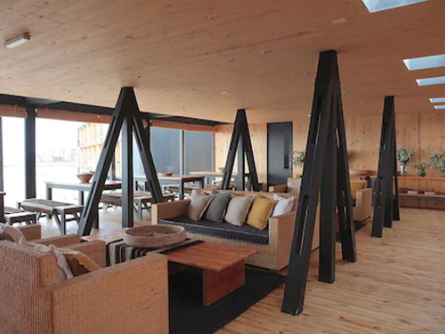 A spacious room with a high ceiling, wooden beams, and large windows. There are several seating areas with cushions and tables.
