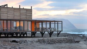A modern beachside building with a wooden exterior and large glass windows, offering views of the ocean and a mountain in the background. - Hotel Apacheta (Arica)