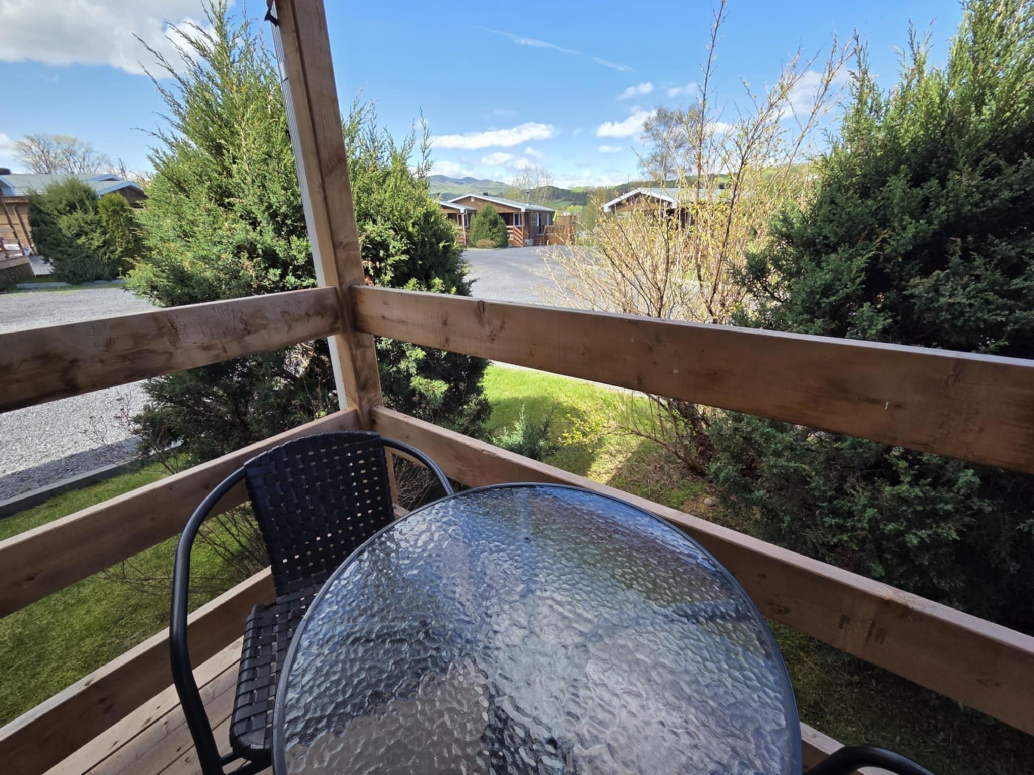 Una terraza de madera con mesa y sillas de vidrio, con vistas a un aparcamiento y edificios en un paisaje verde y montañoso.