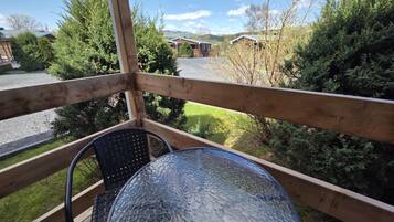 Una terraza de madera con mesa y sillas de vidrio, con vistas a un aparcamiento y edificios en un paisaje verde y montañoso.