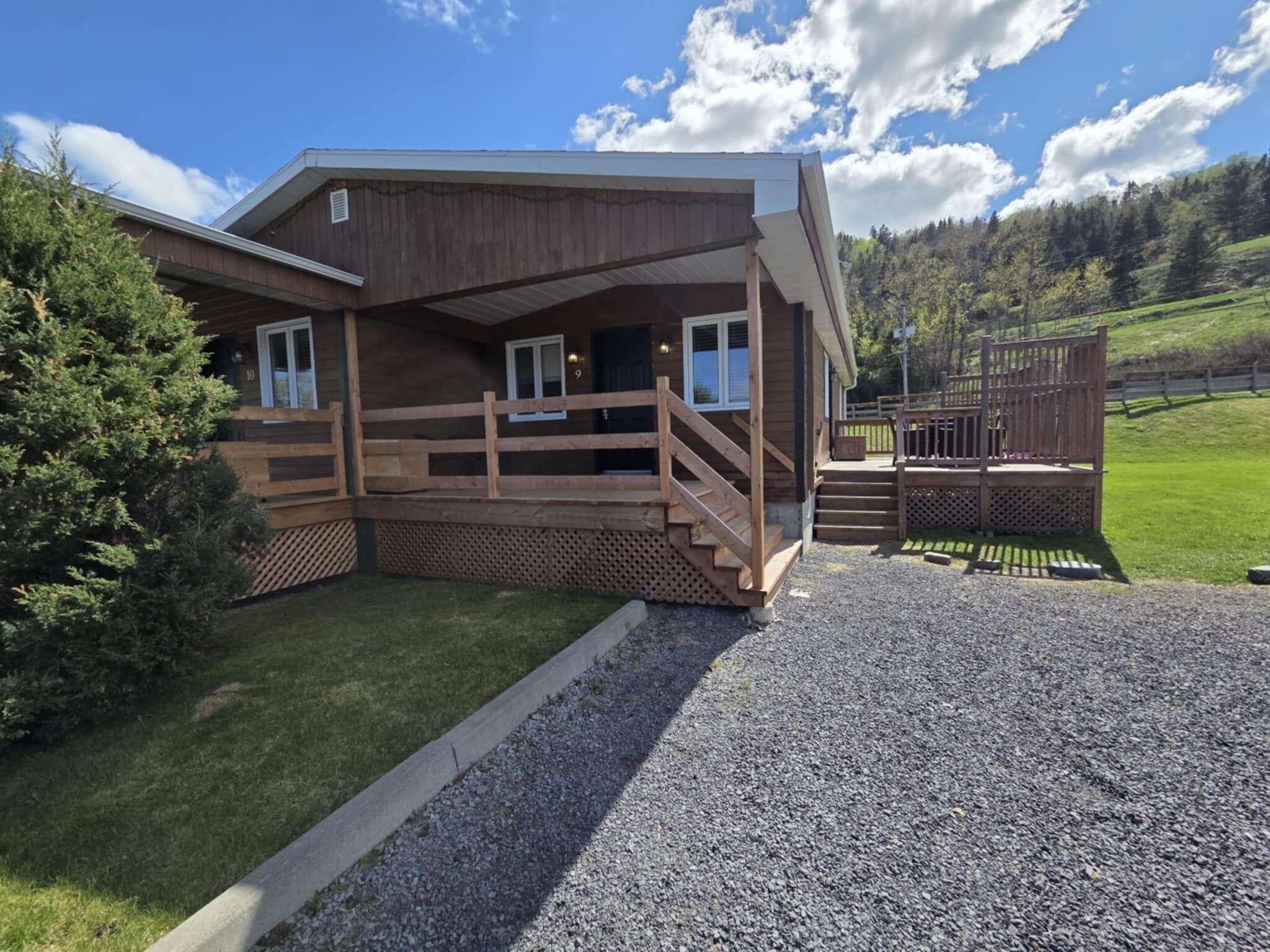 Eine Holzhütte mit Veranda, einem Schotterweg und einem bewaldeten Hang im Hintergrund.