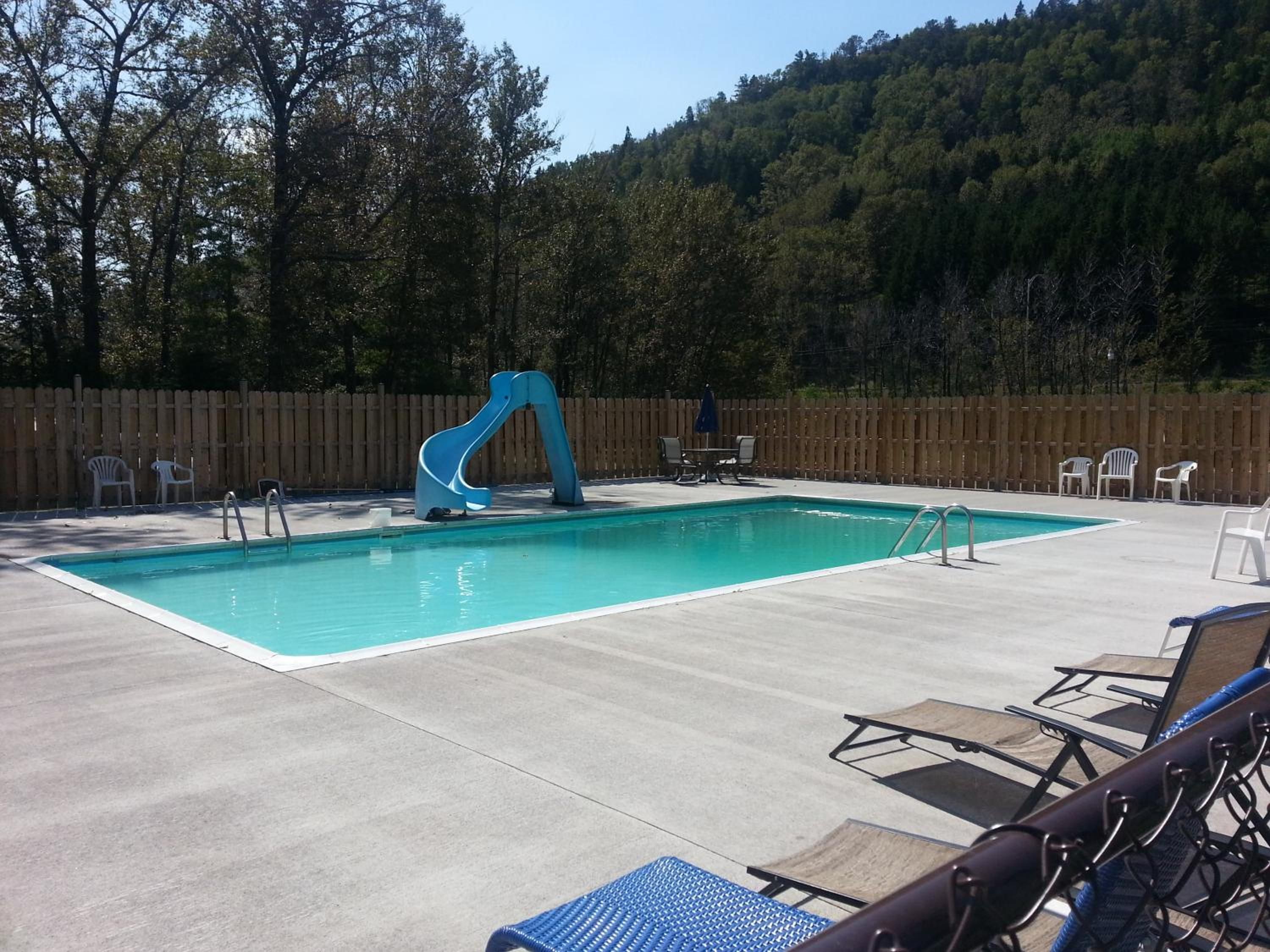 Una piscina rectangular con tobogán, rodeada de sillas de descanso y una valla de madera.