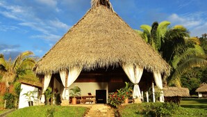 A thatched-roof hut with a staircase leading to the entrance, surrounded by greenery and palm trees. - Sauce Lodge Laguna Azul (Sauce)