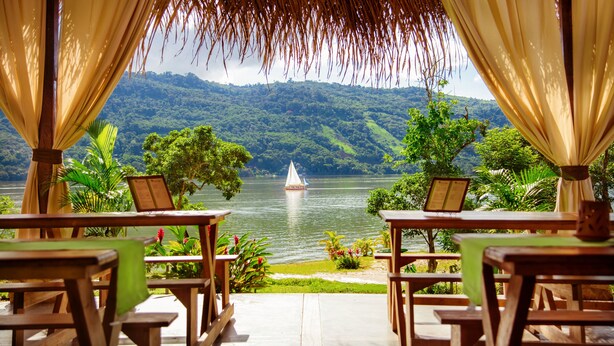 A wooden deck with tables and chairs overlooking a lake with a sailboat, surrounded by lush greenery and hills. - Sauce Lodge Laguna Azul (Sauce)