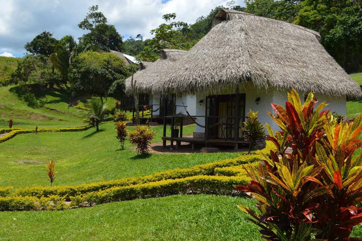 Uma casa com telhado de palha e varanda, rodeada por jardins bem cuidados e uma paisagem acidentada.