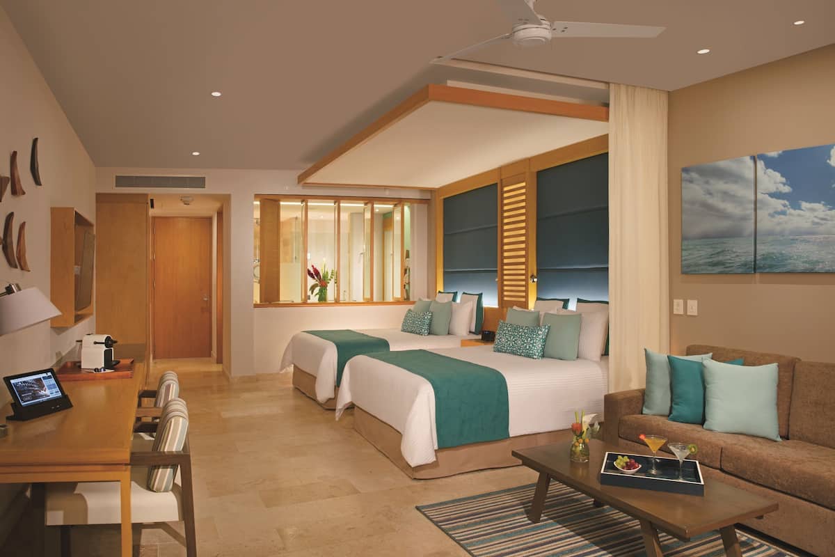 A hotel room with two beds, a sofa, a dining area, and a large window with a view.