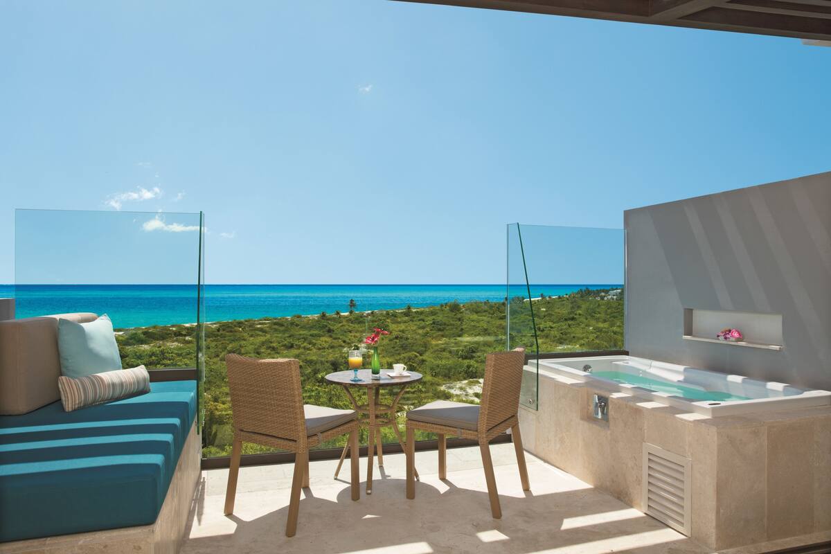 A balcony with a view of the ocean, a seating area with cushions, a table with drinks, and a hot tub.