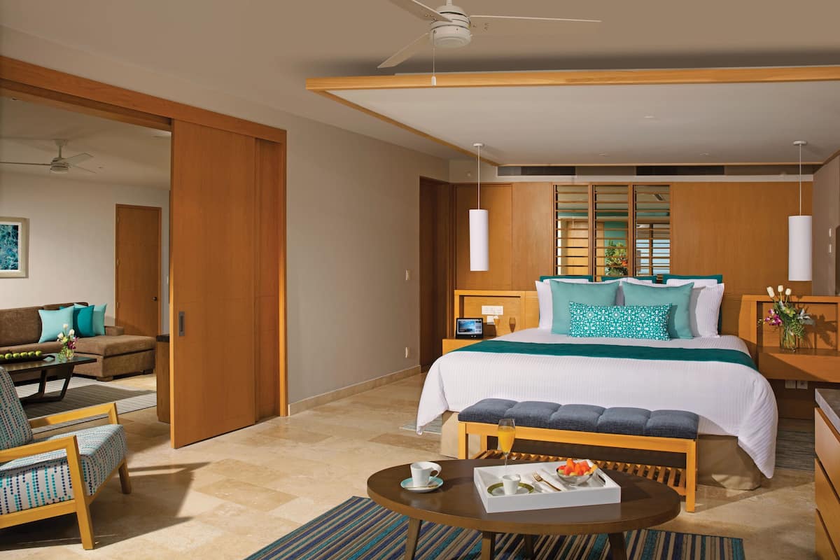 A modern hotel room with a large bed, a sitting area with a sofa and armchairs, a small table with breakfast items, and a view of the outside through large windows.