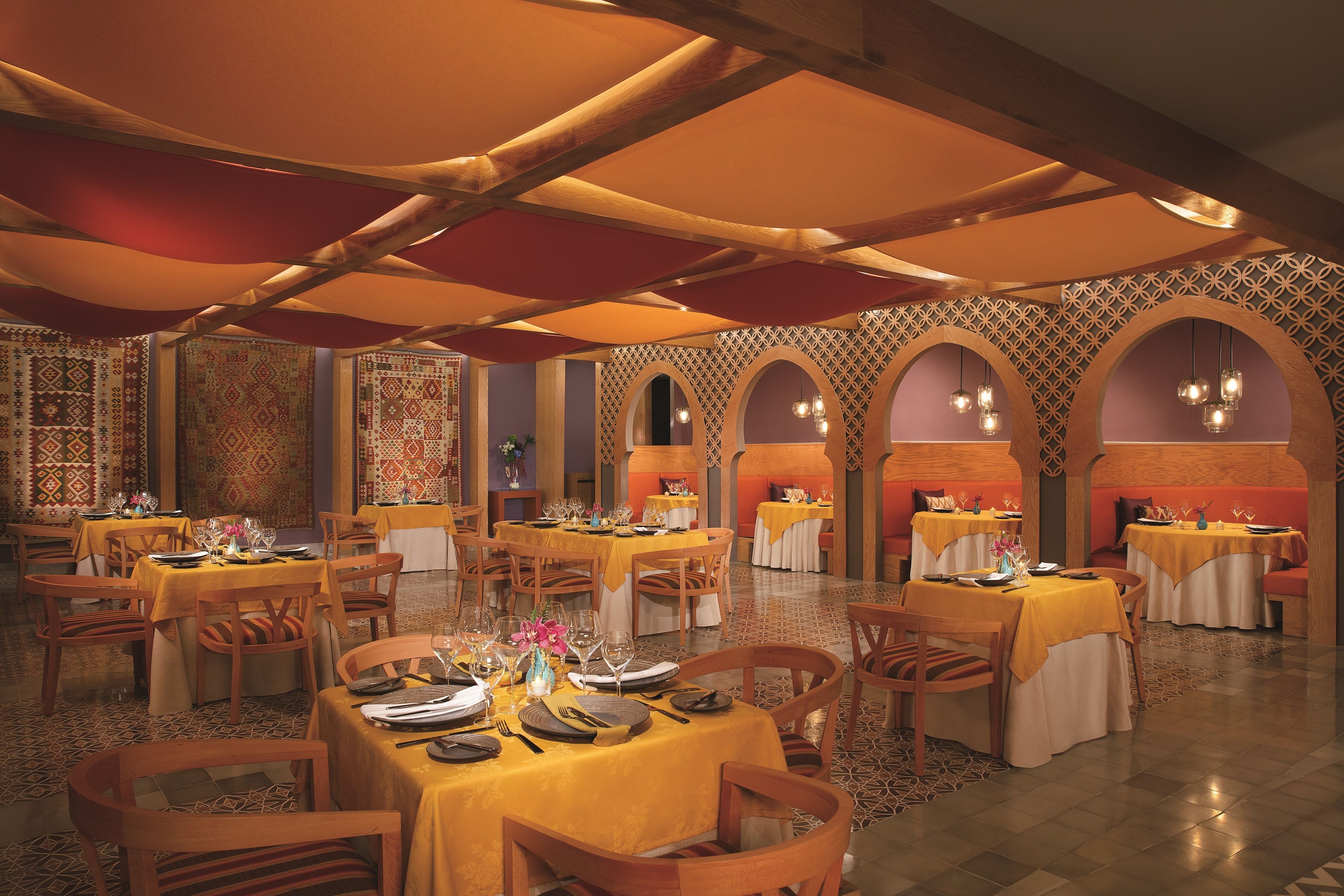 A restaurant with multiple tables, each covered with a yellow tablecloth, and wooden chairs. The room features patterned walls and arches, with hanging light fixtures.