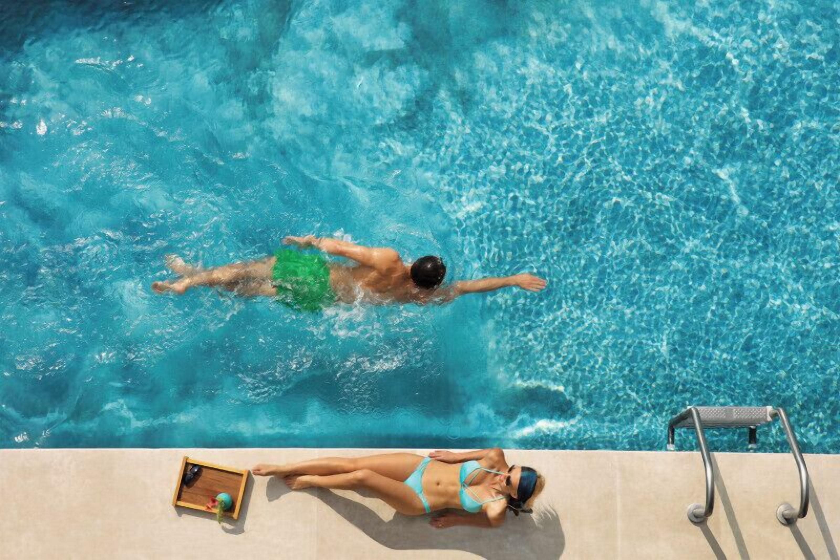A swimming pool with a person swimming and another person sunbathing on the edge.