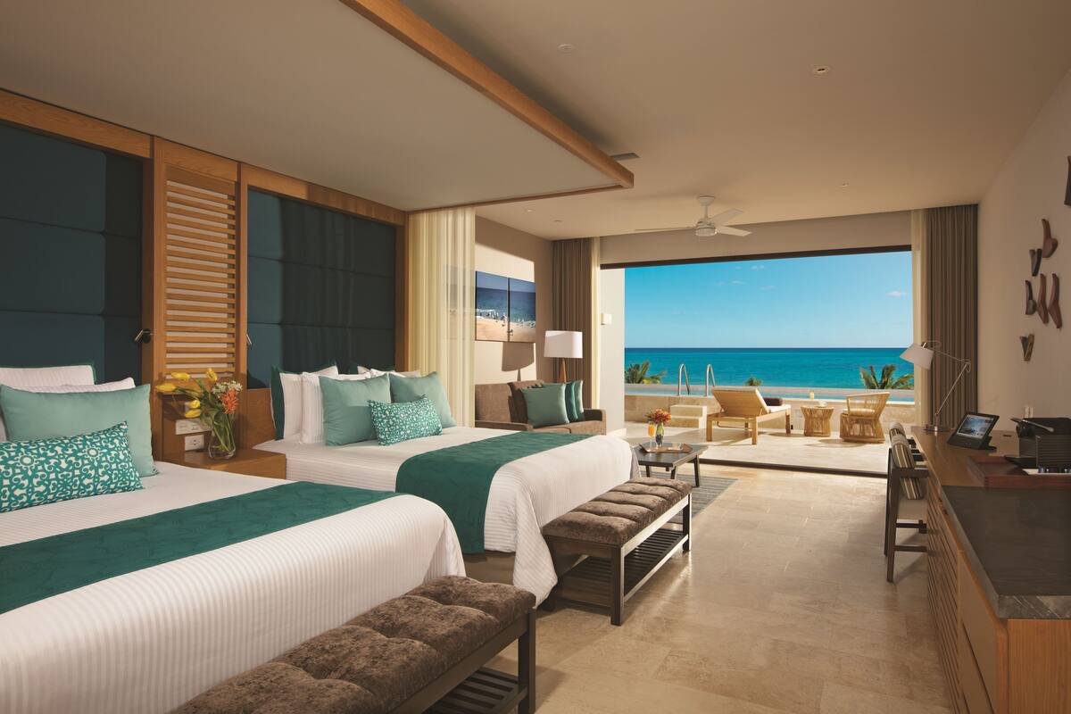 A hotel room with two beds, a sitting area, a desk, and a view of the ocean.