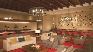 A spacious lobby with a modern design, featuring a seating area with sofas and armchairs, a bar area with red chairs, and a wall with numerous framed pictures.