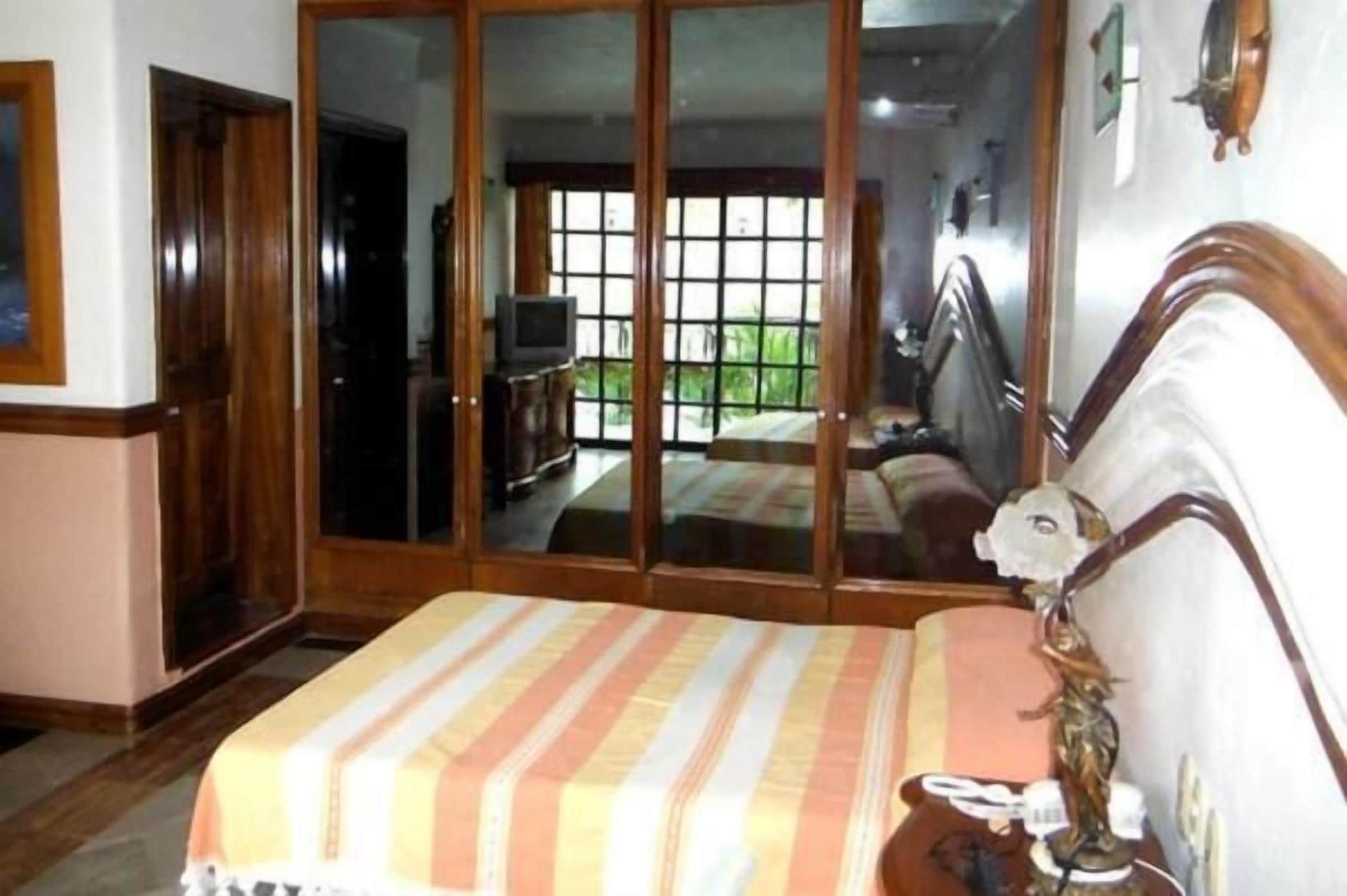 A room with a bed, a wooden wardrobe, a sliding glass door leading to a balcony, and a small table with a lamp.