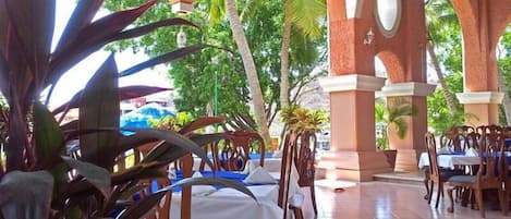 An outdoor dining area with wooden tables and chairs, surrounded by large potted plants and palm trees.