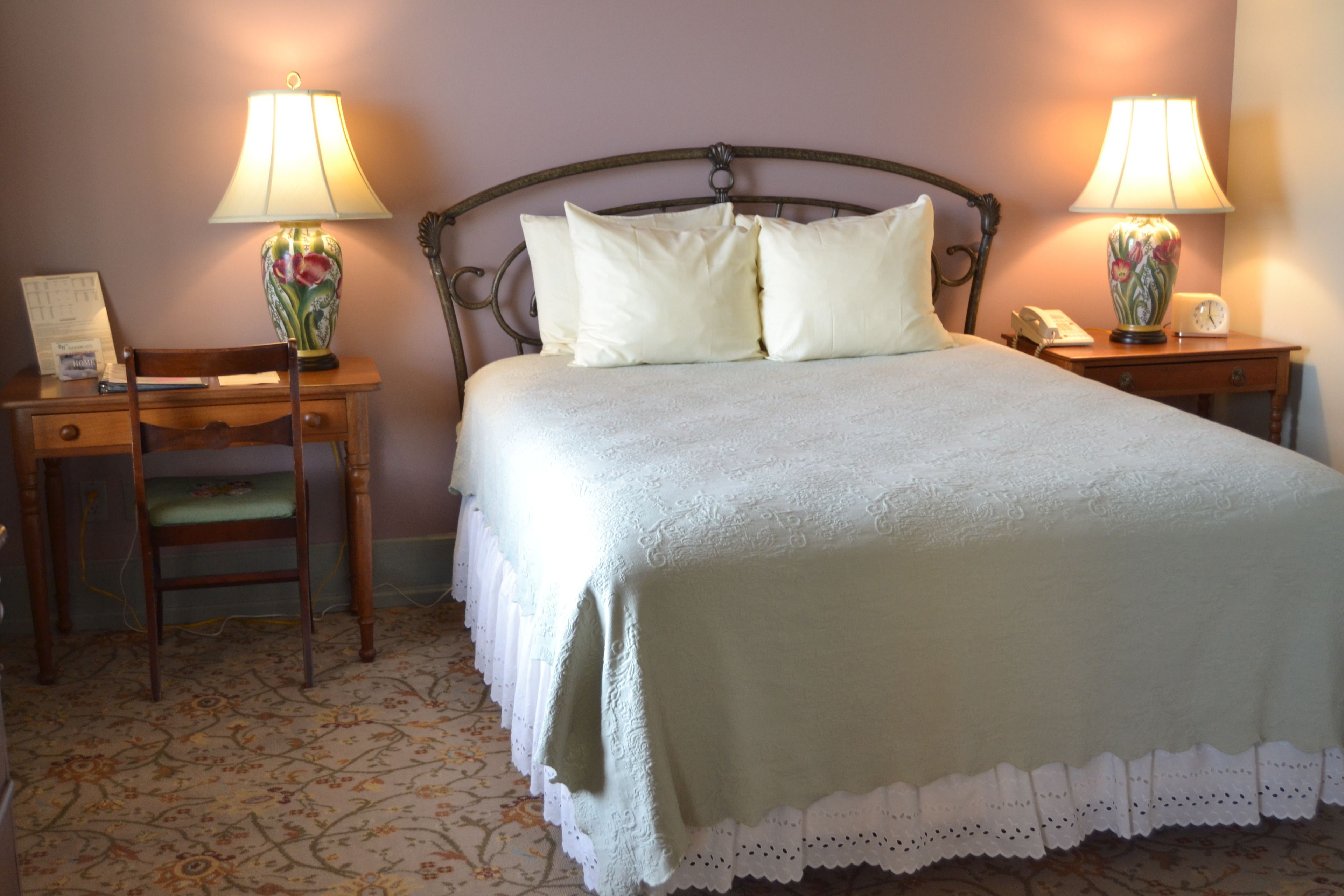 A neatly made bed with a green bedspread and white pillows, flanked by two nightstands with lamps and a chair.