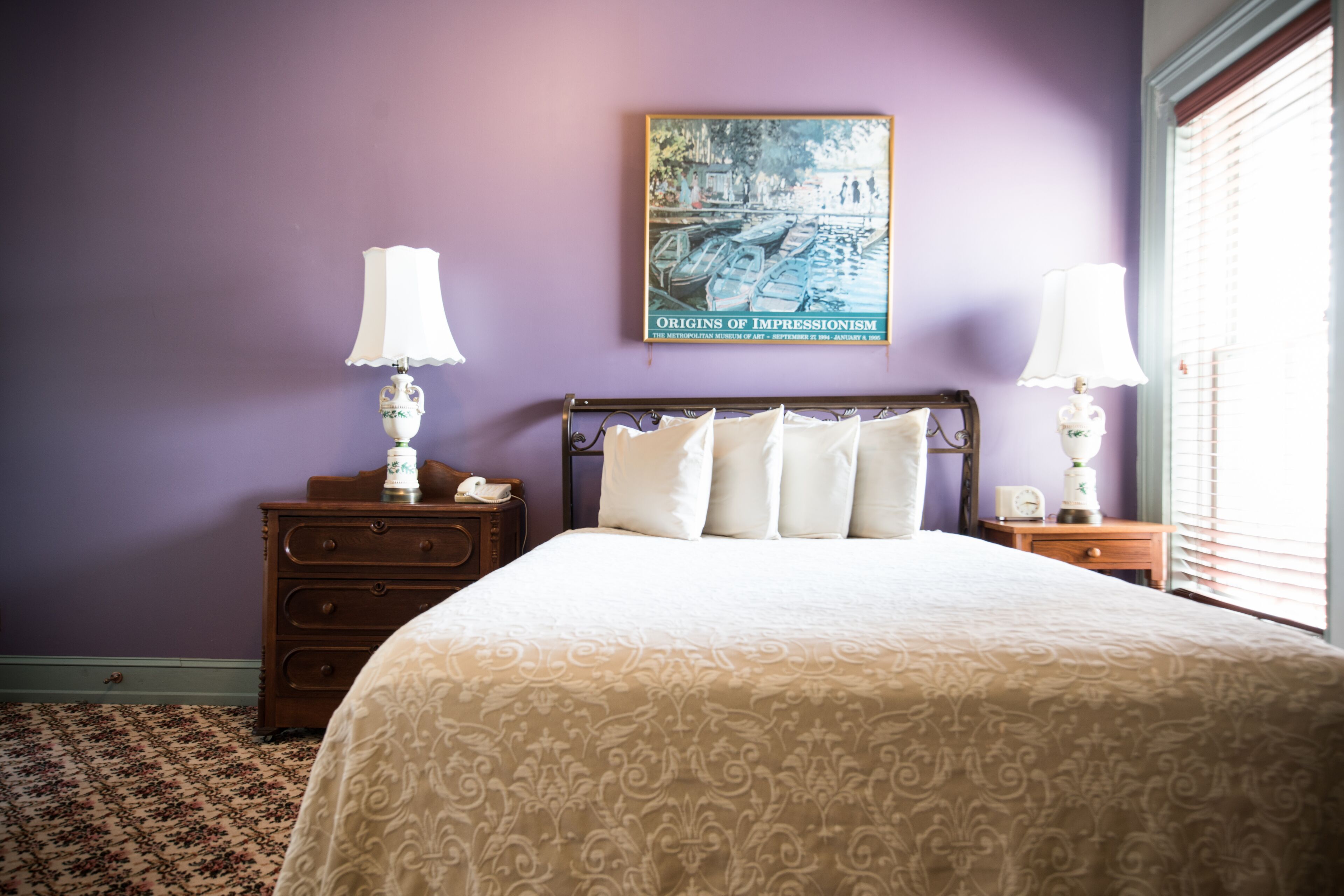 A neatly made bed with a patterned bedspread, two bedside tables with lamps, a wall-mounted painting, and a window with blinds.