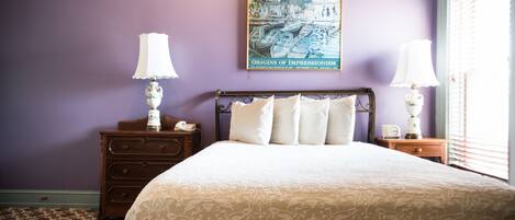 A neatly made bed with a patterned bedspread, two bedside tables with lamps, a wall-mounted painting, and a window with blinds.
