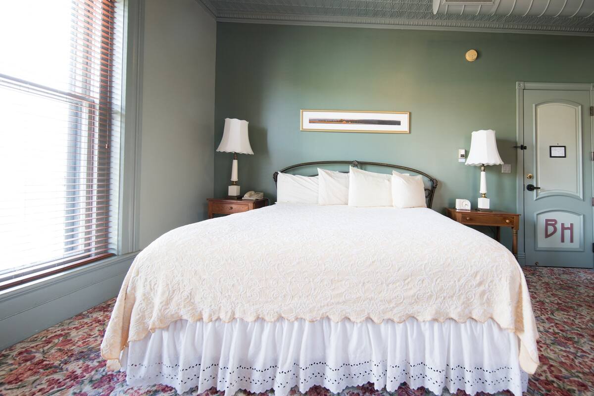 A neatly made bed with a white lace bedspread and a floral-patterned skirt, two bedside tables with lamps, a framed picture on the wall, and a door with a 'BH' monogram.
