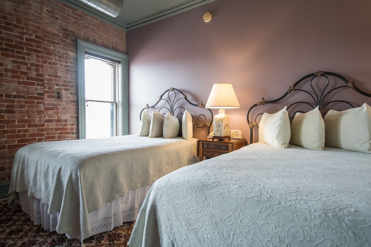 A room with two beds, a brick wall, a window, and a nightstand with a lamp.
