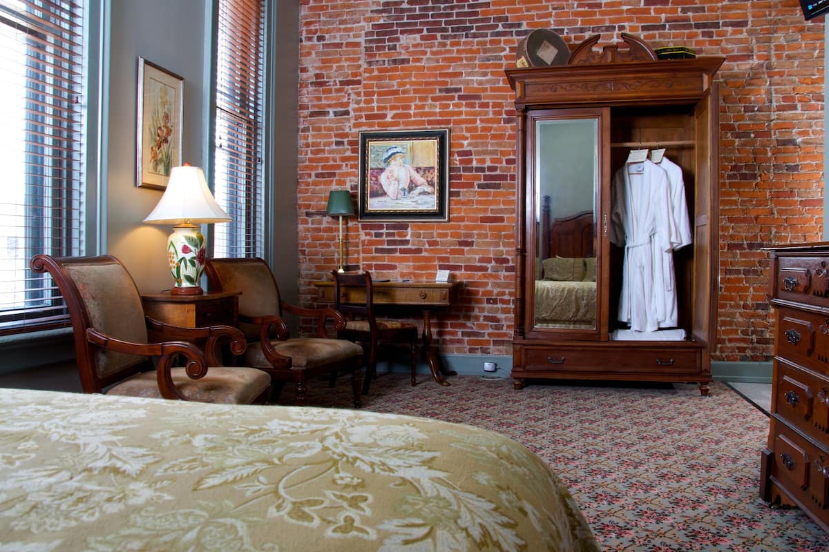 A hotel room with a brick wall, a wooden wardrobe, a bed with a floral bedspread, a desk with a lamp, and a chair.