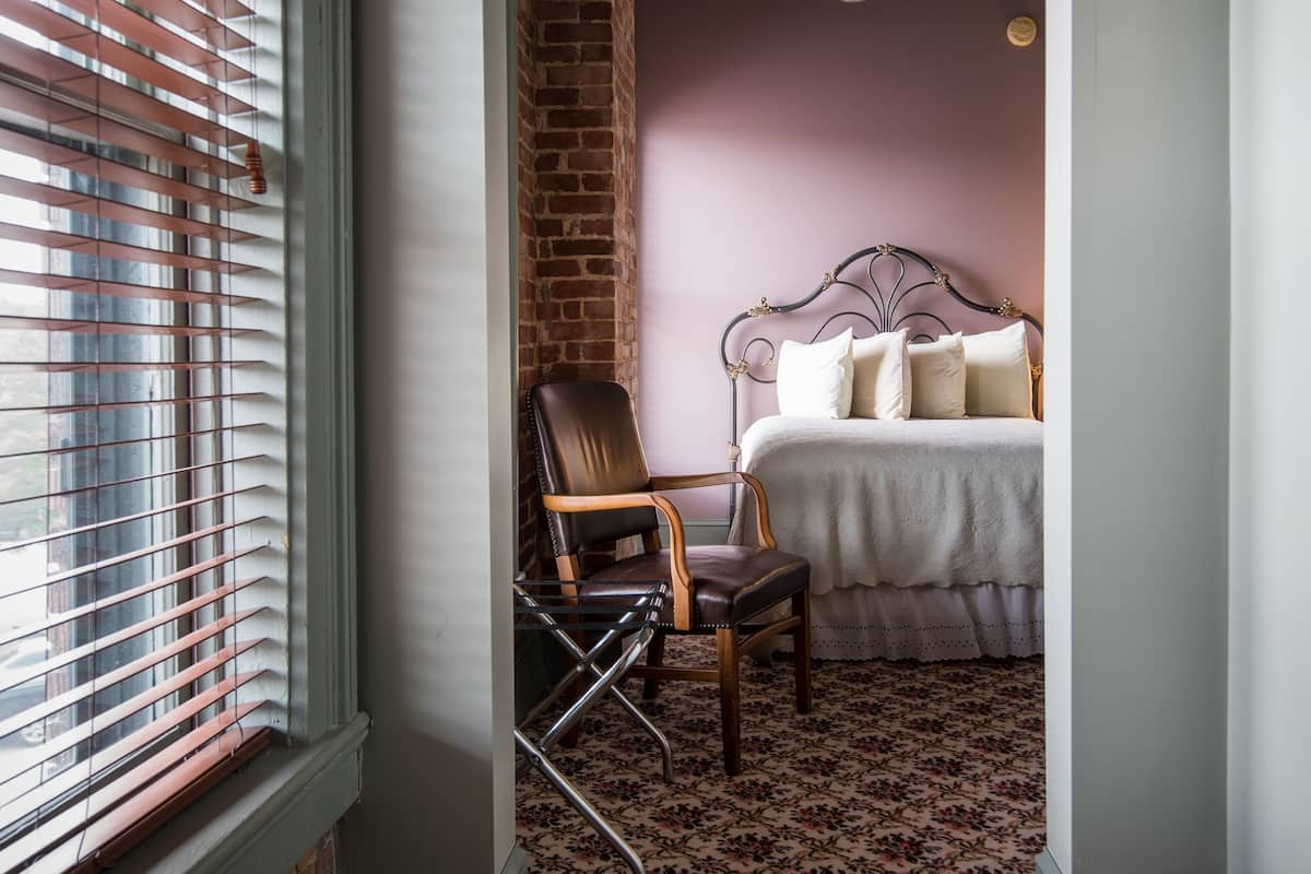 A narrow hallway leading to a bedroom with a bed, a chair, and a patterned carpet.