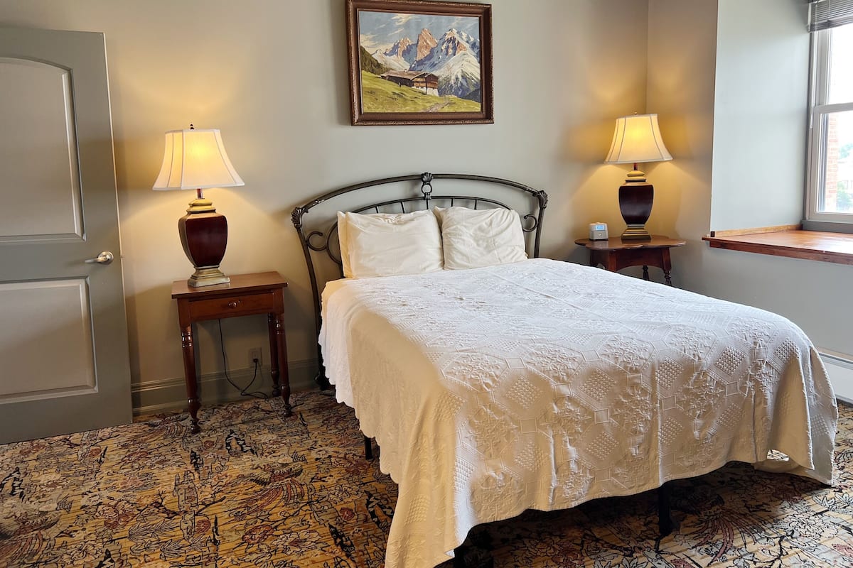 A neatly arranged bedroom with a bed, two bedside tables, a painting on the wall, and a window with a view.