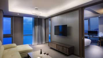 A modern hotel room with a large TV, a sofa, a dining table with wine, and a view of the ocean.