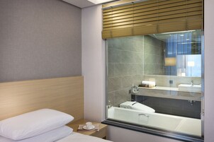 A modern hotel bathroom with a glass shower enclosure, a visible sink, and a neatly made bed with pillows.