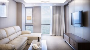 Ein modernes Hotelzimmer mit einer großen Couch, einem Flachbildfernseher, einem Esstisch mit einer Flasche und Gläsern sowie Meerblick durch große Fenster.