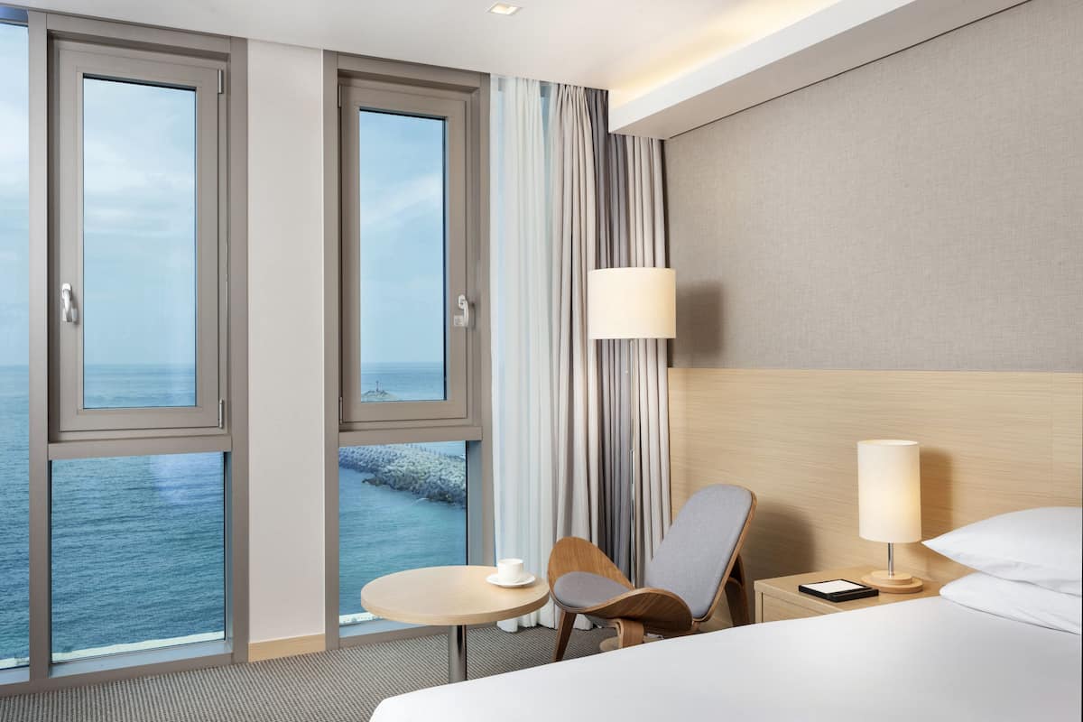 Une chambre d’hôtel avec un lit, une table de chevet avec une lampe, une chaise, une petite table avec une tasse, et une vue sur la mer par de grandes fenêtres.