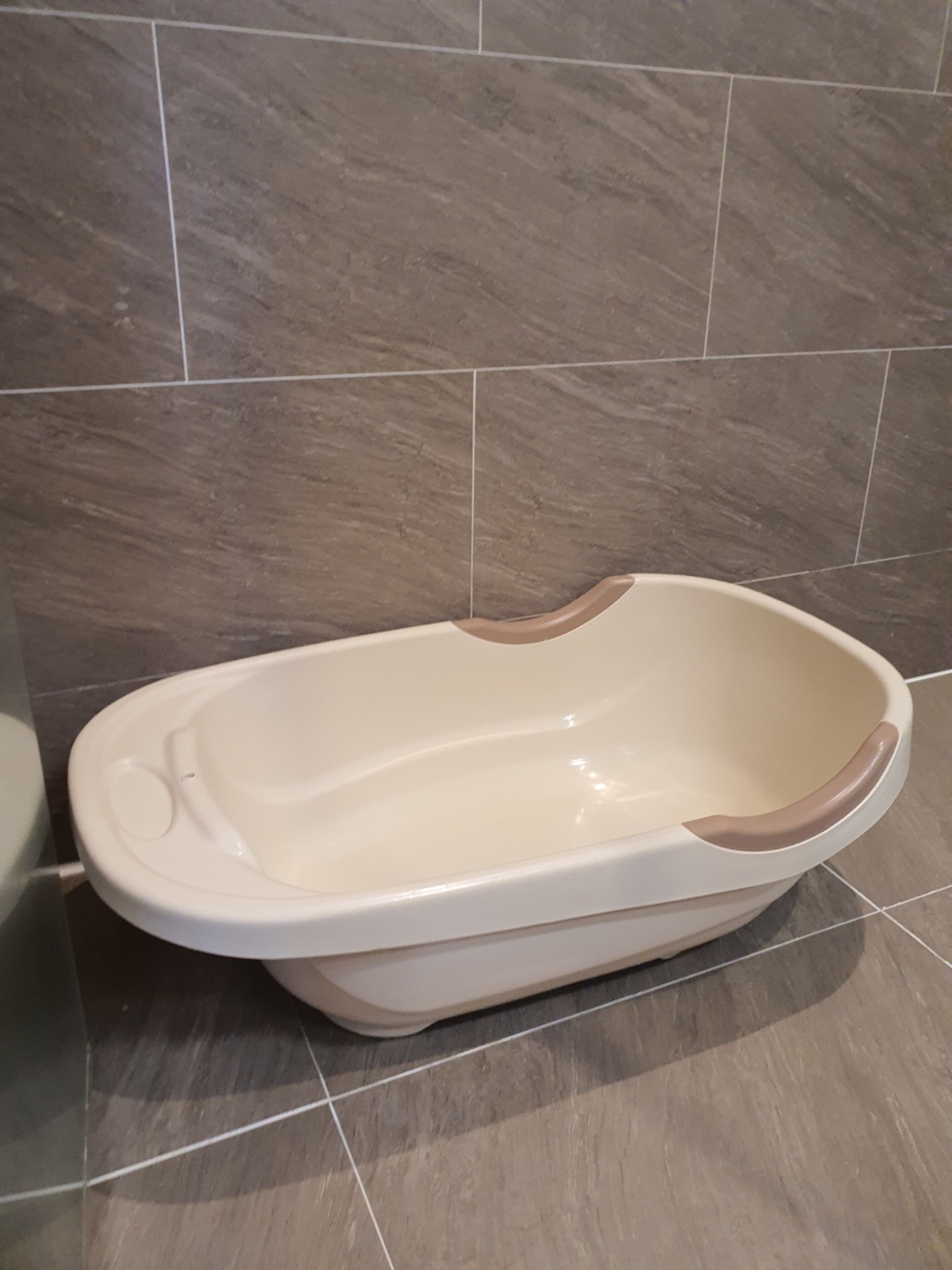 A white baby bathtub with a brown edge in a bathroom with grey tiled walls and floor.