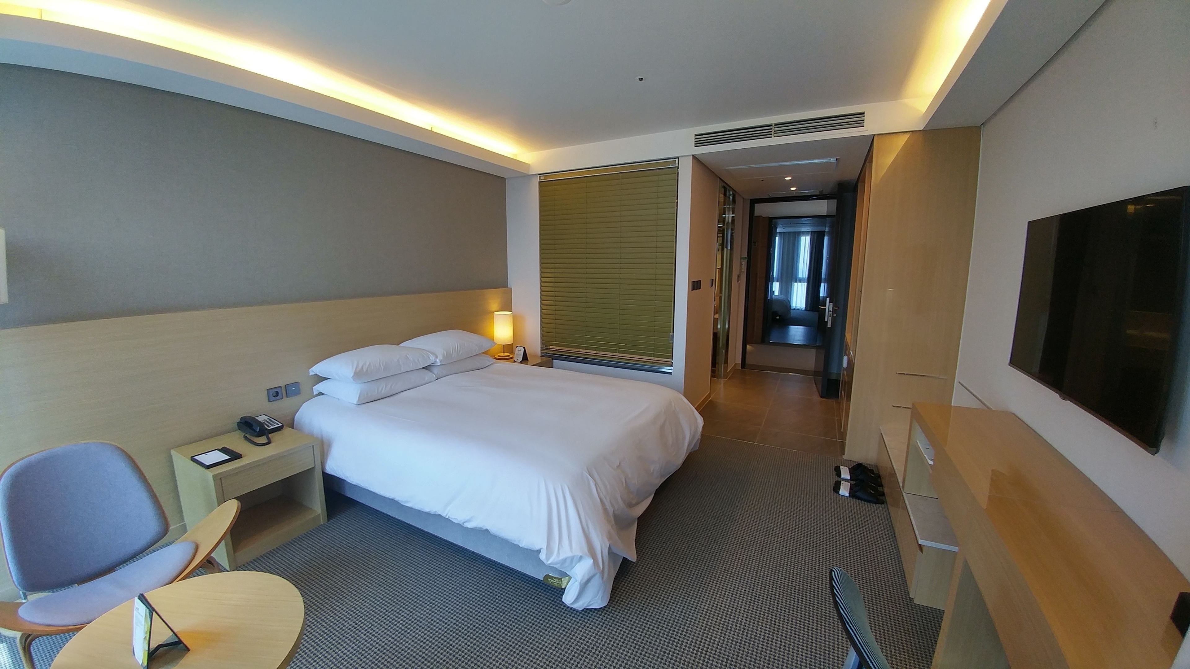 Ein Hotelzimmer mit Bett, Nachttisch, Lampe, Sessel und einem an der Wand befestigten Fernseher.