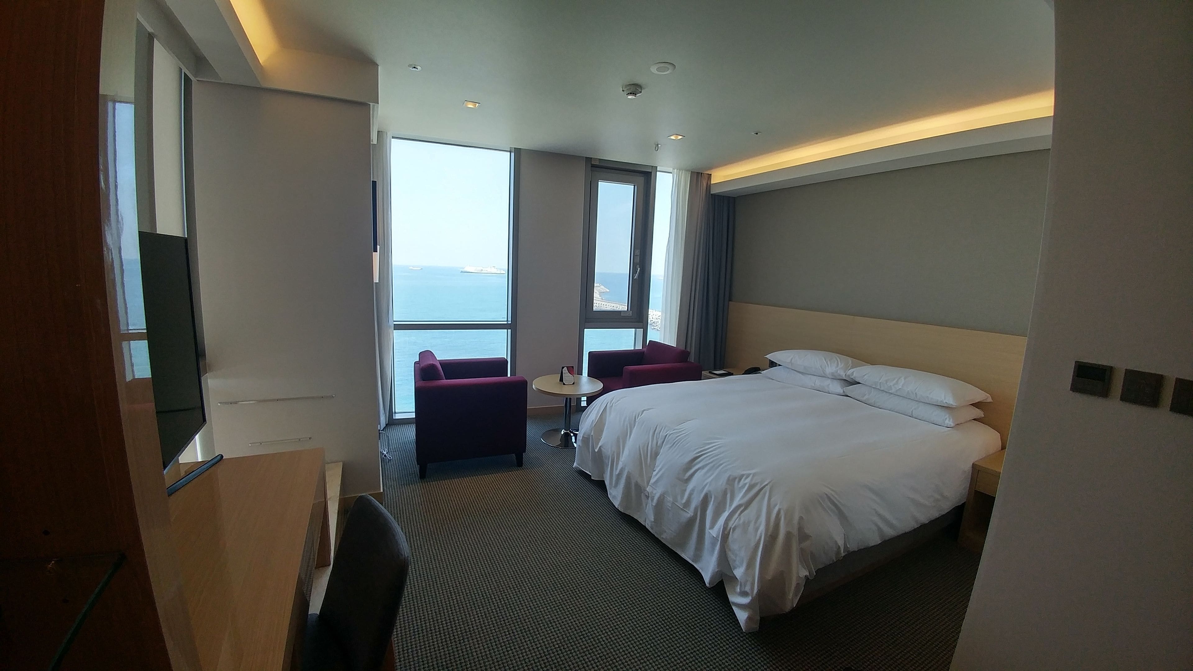 A hotel room with a large bed, a desk, a chair, and a small table. There are large windows with a view of the sea.