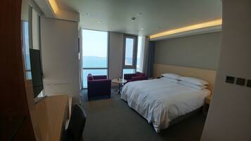 A hotel room with a large bed, a desk, a chair, and a small table. There are large windows with a view of the sea.