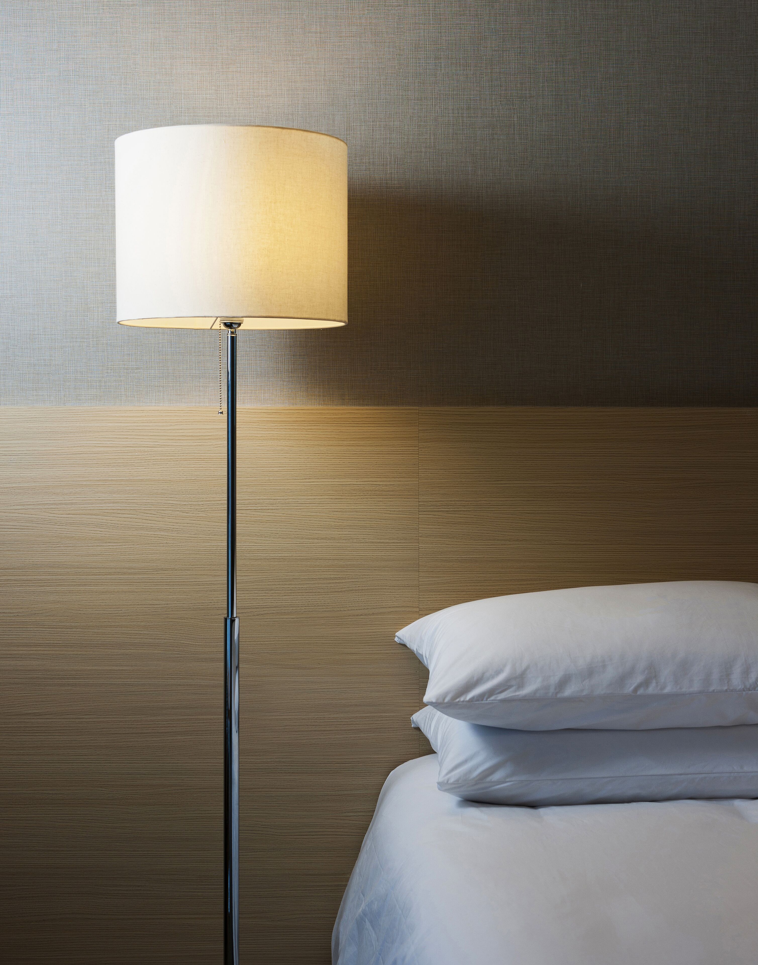 A modern hotel room with a wooden headboard, a floor lamp, and white bedding.
