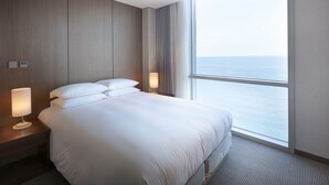 Ein Hotelzimmer mit einem großen Bett, zwei Nachttischlampen, einem Nachttisch und Meerblick durch ein großes Fenster.