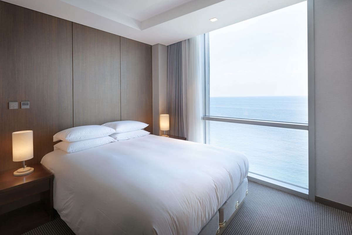 Une chambre d’hôtel avec un grand lit, deux lampes de chevet, une table de nuit et une vue sur la mer par une grande fenêtre.