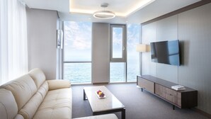 Ein modernes Hotelzimmer mit einer Couch, einem Couchtisch mit Obst, einem Fernseher und einem großen Fenster mit Meerblick.