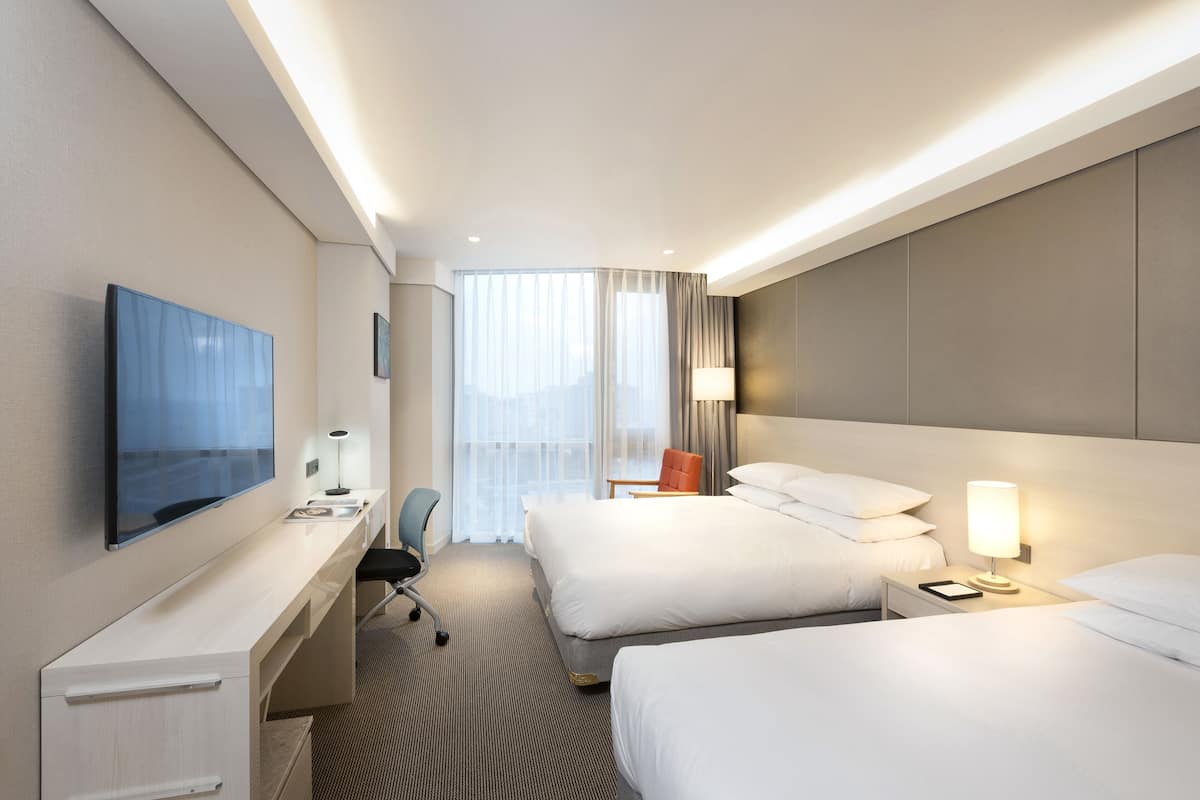 Une chambre d’hôtel avec deux lits, un bureau doté d’un téléviseur à écran plat, une chaise, une table de chevet avec une lampe et une grande fenêtre avec des rideaux.