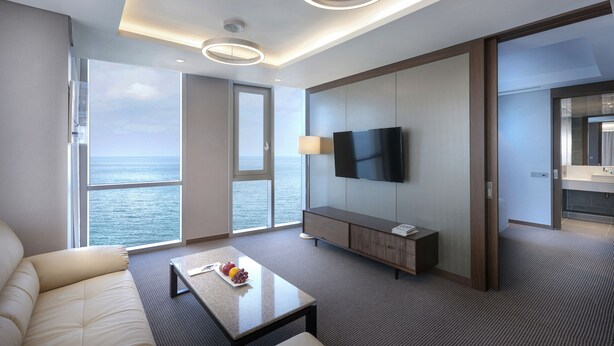 Ein modernes Hotelzimmer mit großem Fenster, Flachbildfernseher, Sofa, Couchtisch mit Früchten und einem Holzschrank.