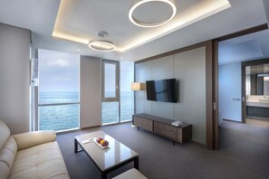 Una habitación de hotel moderna con un ventanal grande, un televisor de pantalla plana, un sofá, una mesa de centro con fruta y un armario de madera.